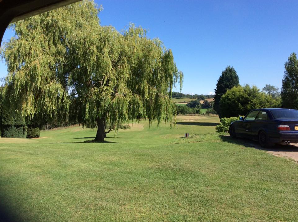 A blue car is parked on the edge of Bank farms pitch and put golf course. The ground falls away and in between the courses greens are a number of trees, a large Willow is central with sculptured Fir trees to the left and right. Beyond the course is a spectacular countryside view down the Severn Valley.